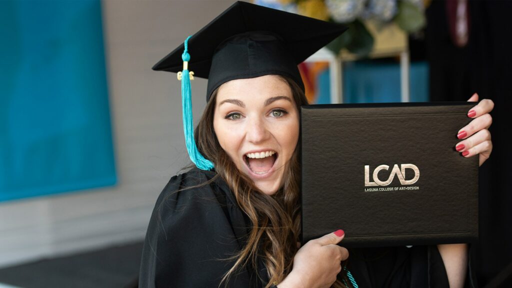 A Student with long brown hair in a graduation cap smiles while holding up their LCAD diploma.